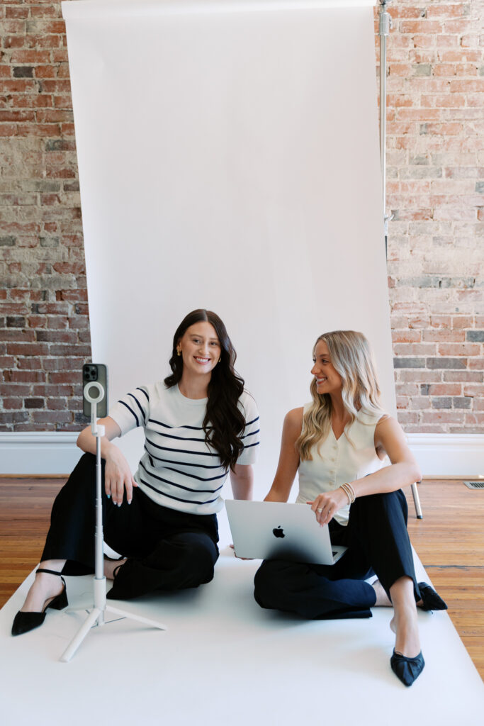 Two social media team members seated on a white backdrop with a laptop and phone tripod