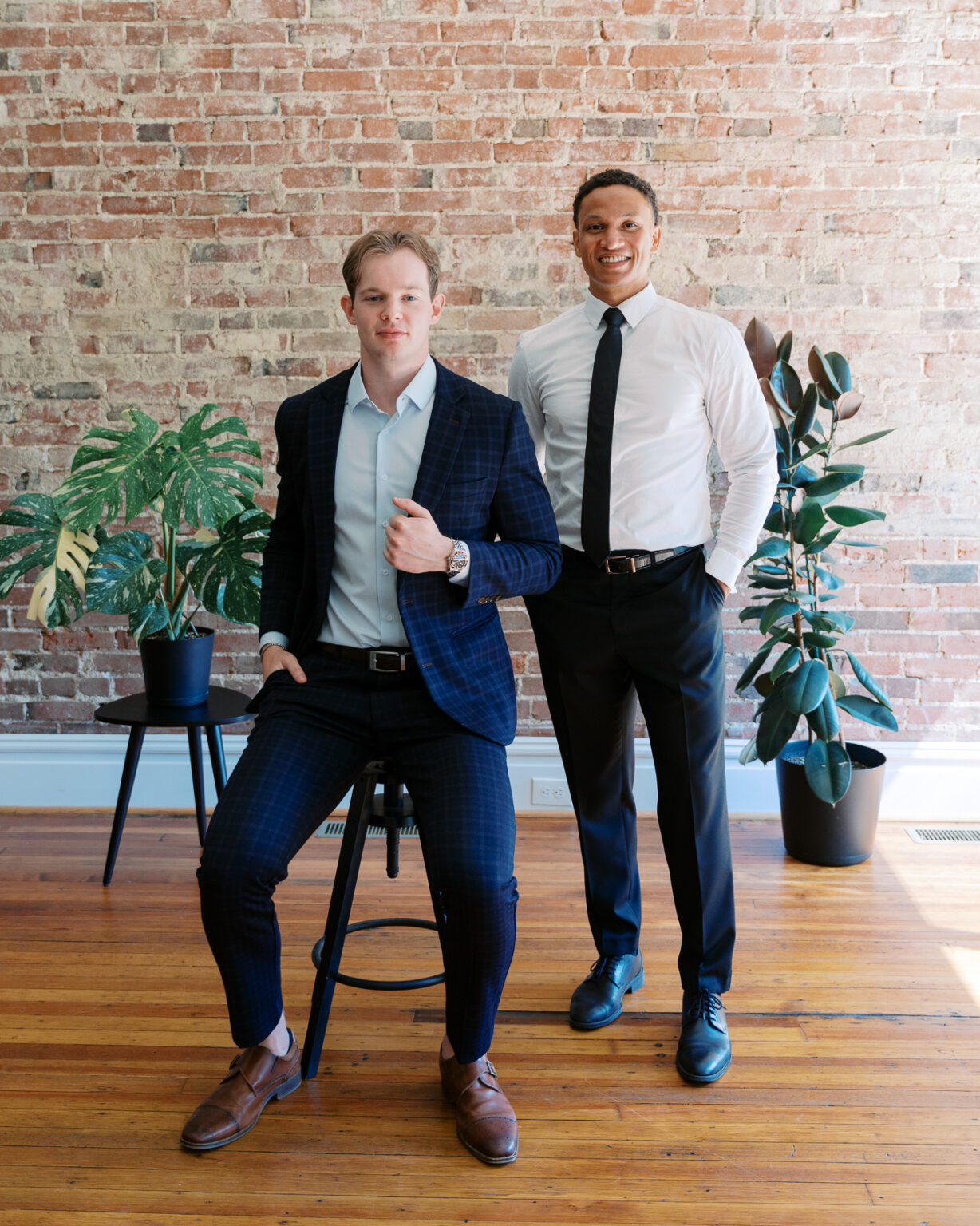 Two team members in business attire posing against an exposed brick wall with plants